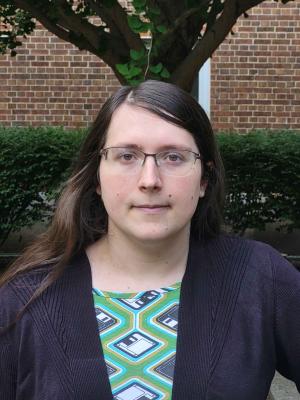 A 'headshot'-style photograph of Alexis. She is wearing a green dress with floppy disks printed on it, as well as a black sweater. She is standing in a verdant environment, with a tree and a brick wall in the background.